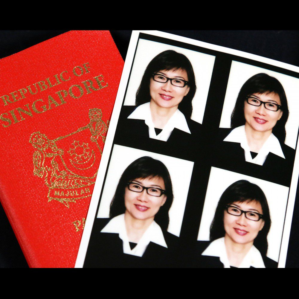 A Singapore passport next to a sheet of four passport-sized photos of a woman wearing glasses and a black suit with a white collared shirt.
